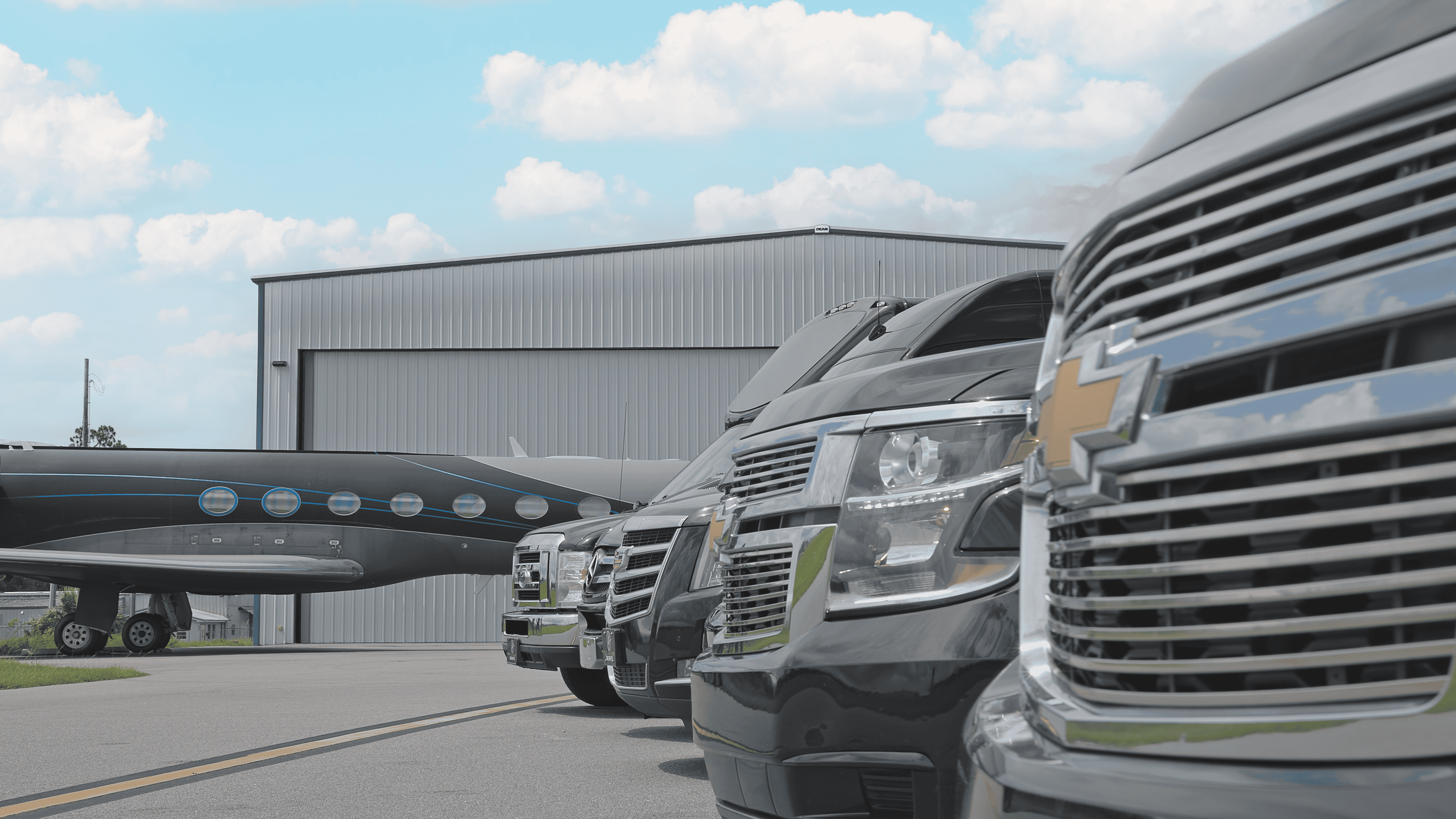 Fleet of Chevrolet Suburban SUVs and executive vehicles parked beside a private jet hangar, providing luxury airport black car service.