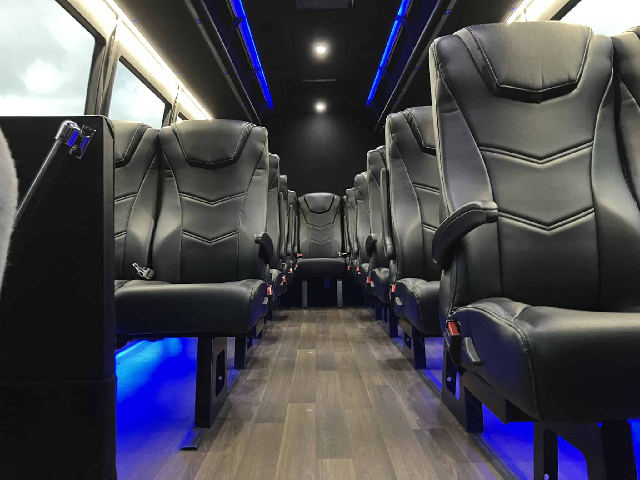 Interior of a luxury 24-passenger executive shuttle bus with reclining leather captain's chairs, center aisle, hardwood flooring, and ambient LED lighting.